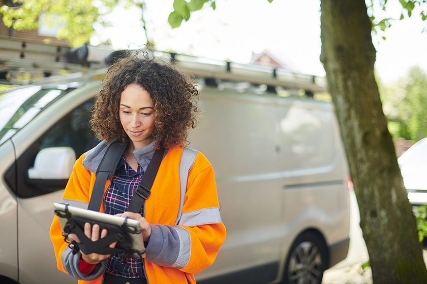 A woman wearing high-vis and holding an iPad stands in front of her work van. 